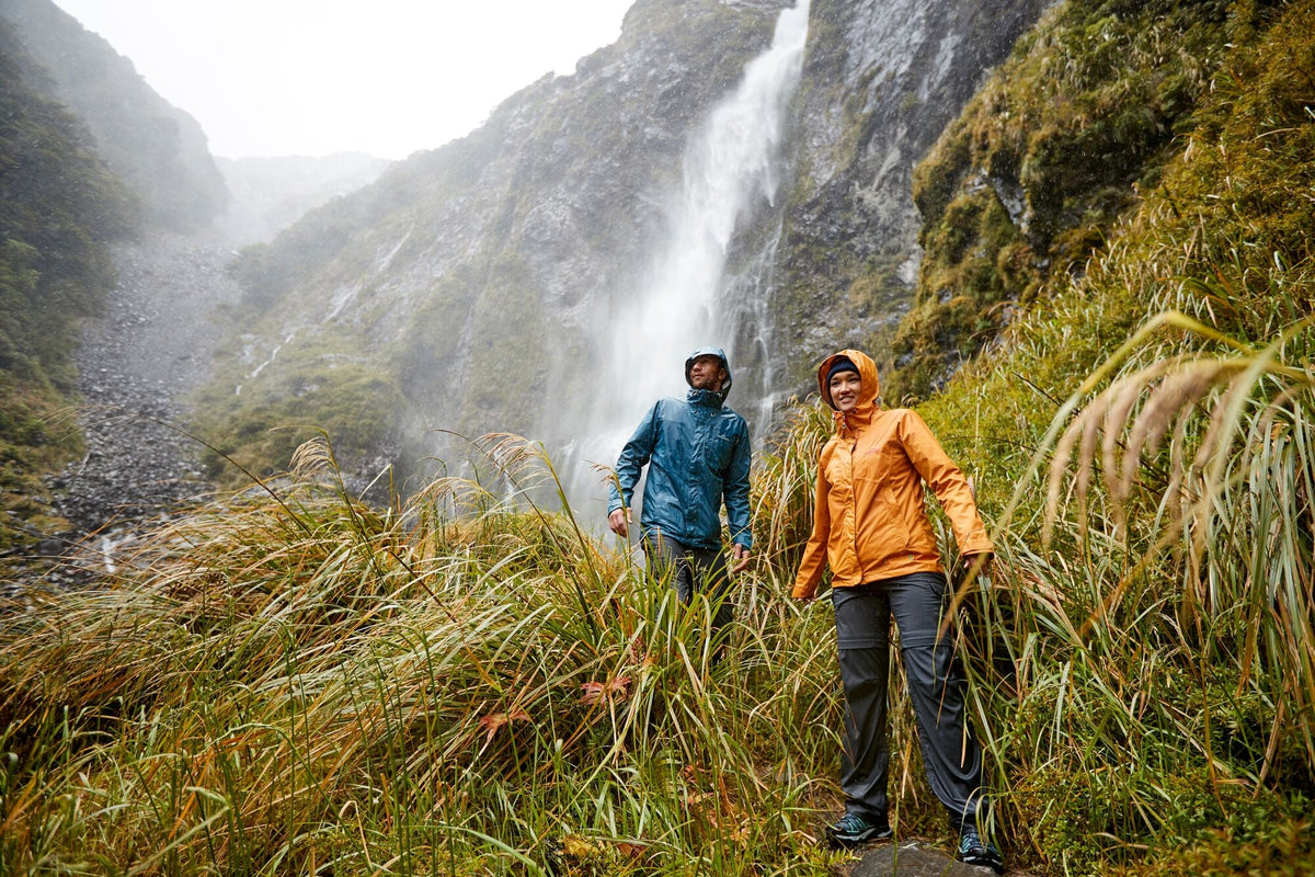 A couple walking through wet long grass in front of a waterfall.